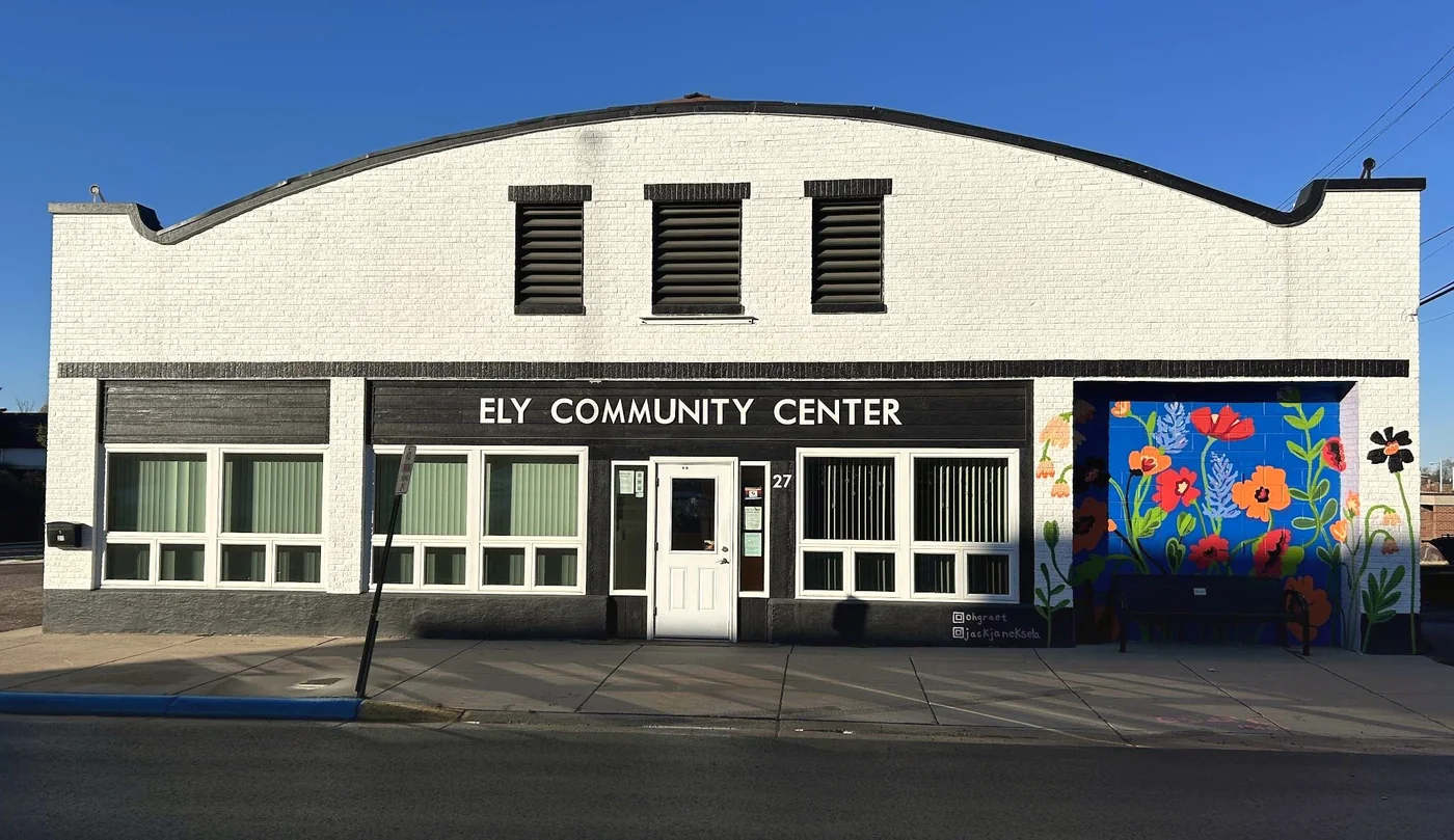 The Ely Community Center in Ely, Minnesota: a white-painted brick building with an arched roofline, a black sign band reading ELY COMMUNITY CENTER, and a bright flower mural on a blue wall at right